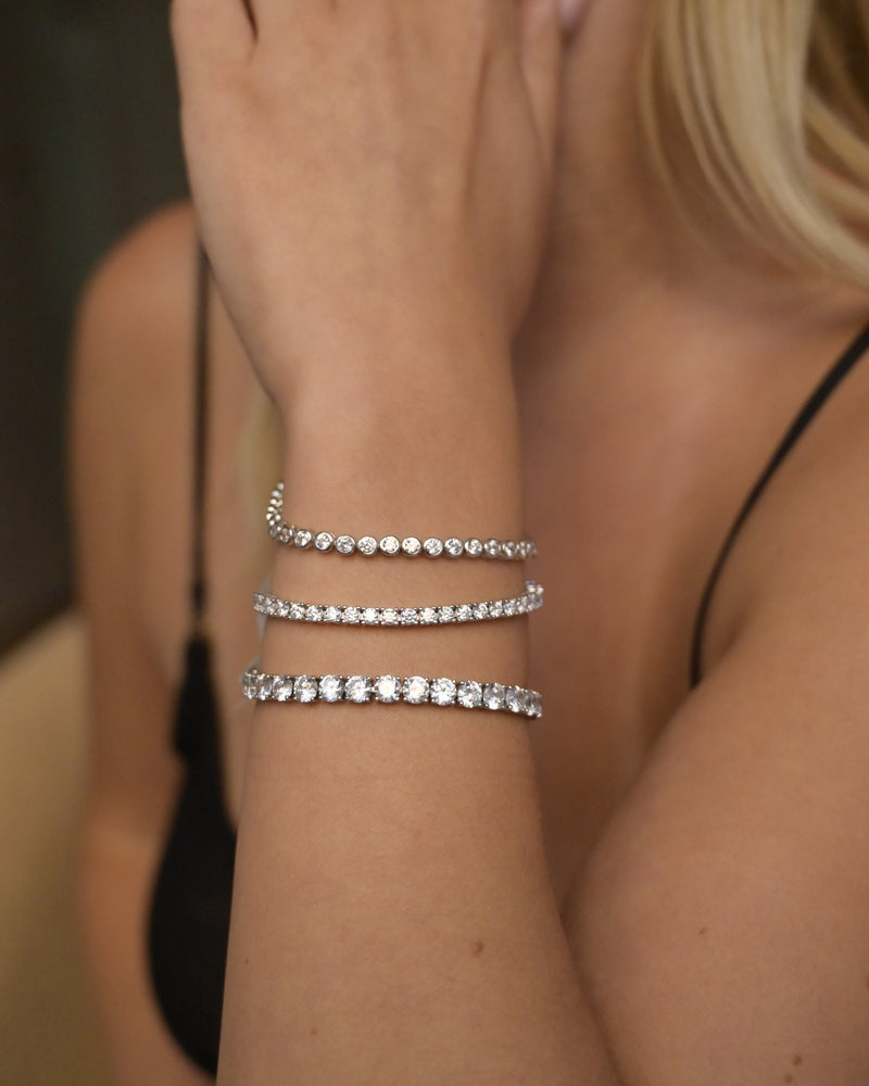 A close-up of a woman's wrist wearing three thin silver tennis bracelets with sparkling clear stones, shown against her skin and black top.